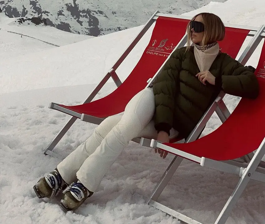A woman sitting on red foldable chairs posing with skiing equipment.